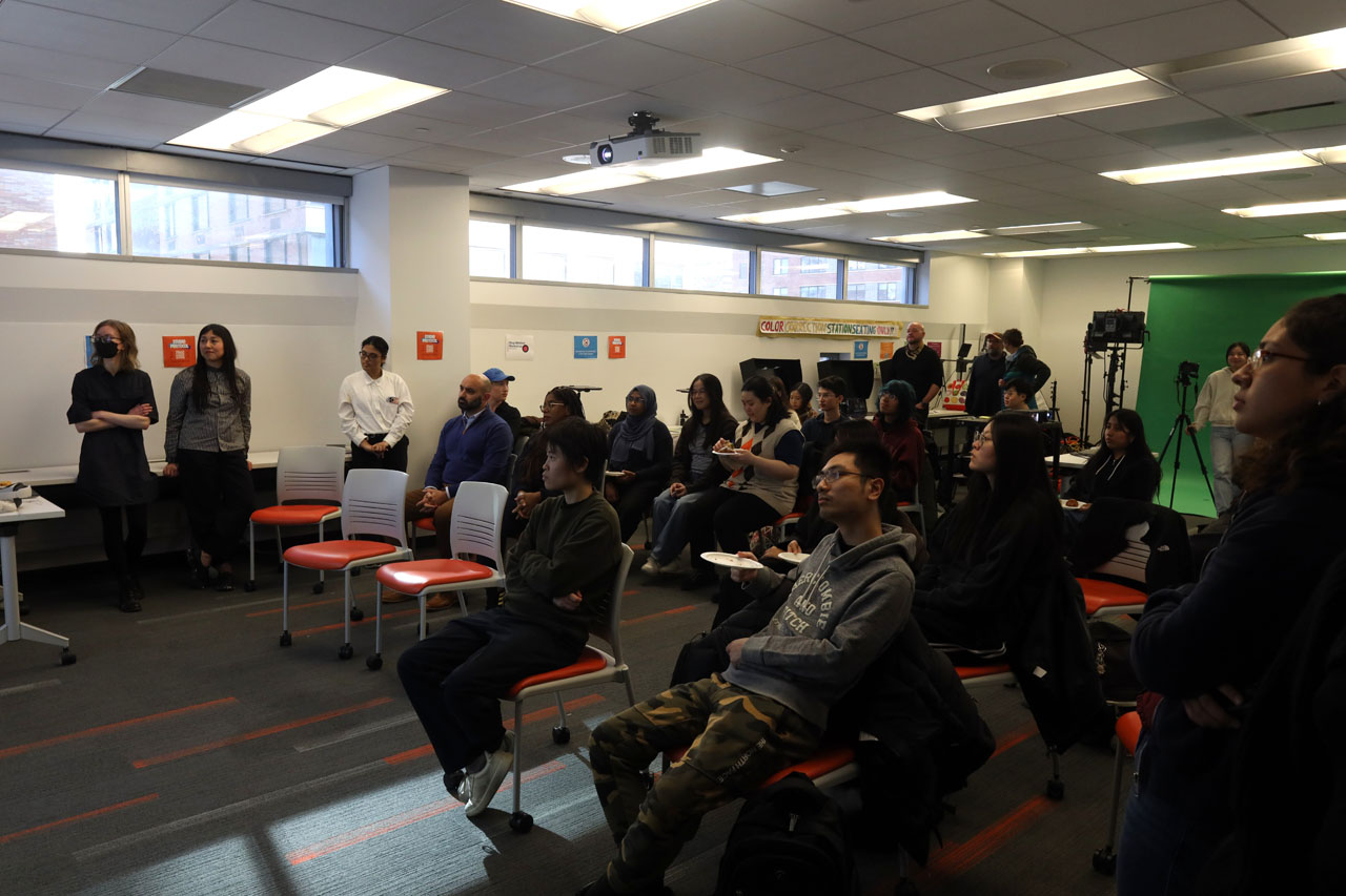 Visitors sitting in rows of orange rolling chairs watching a presentation in the NMA Studio. A filming set-up with green screen is set up in the back of the room.