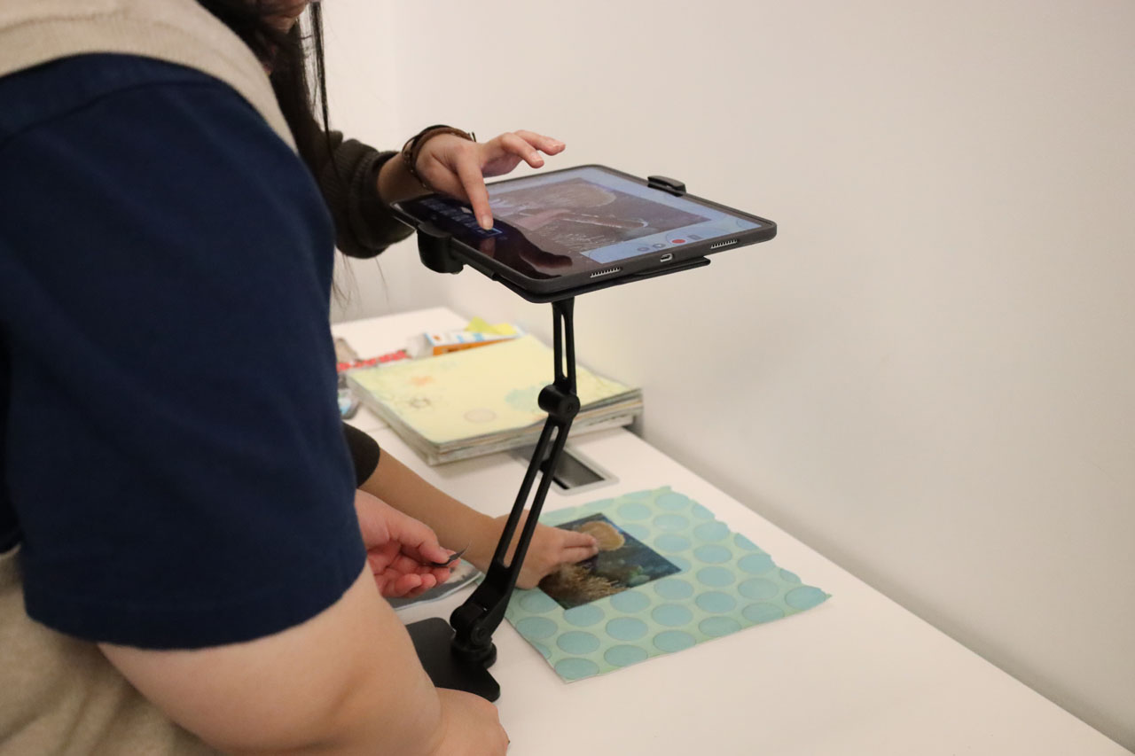 Two students use an iPad on a stand to create a stop-motion animation with collaged materials.