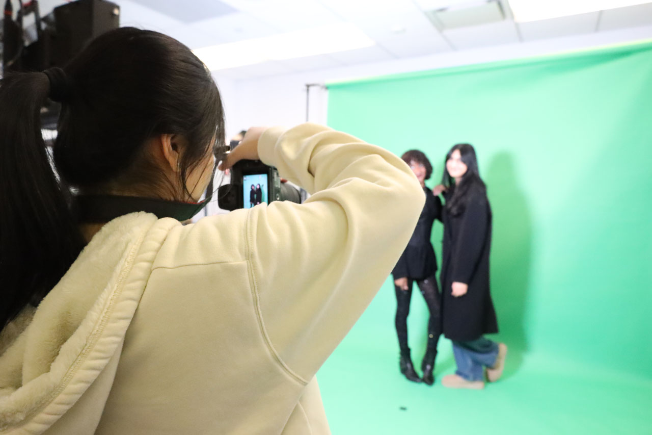 A student takes a photo of two other students posing against a green screen.