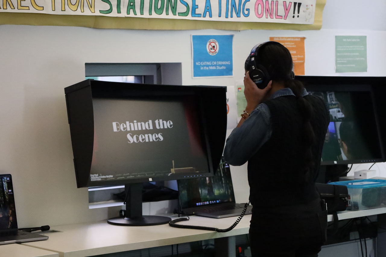 A student wearing a headset watches a screen showing student work. The screen reads 'Behind the Scenes' in a Broadway font.