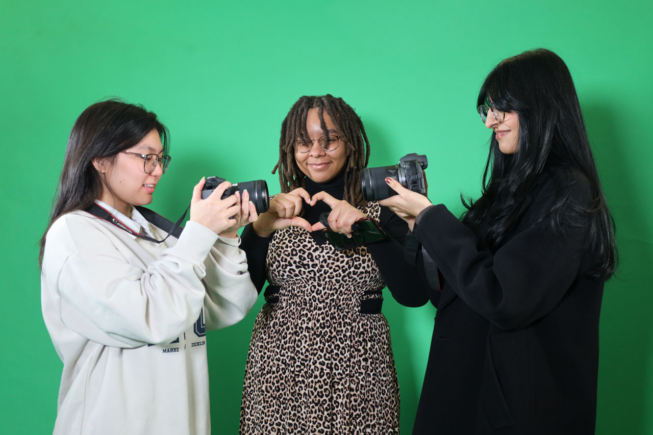 Three students stand against a green screen, facing each other, with the two outer students holding up cameras, looking at each other, and the middle one making a heart with their hands, facing the camera.
