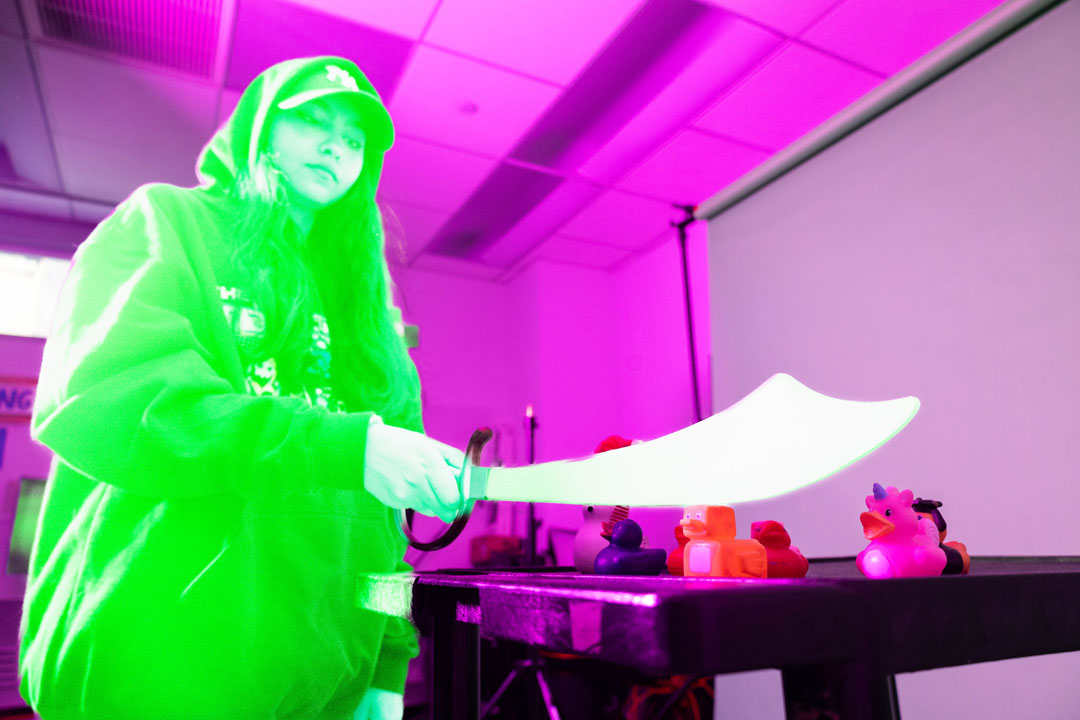 A student holding a prop sword up to the camera. The figure is edited to be neon green, and the background is pink.