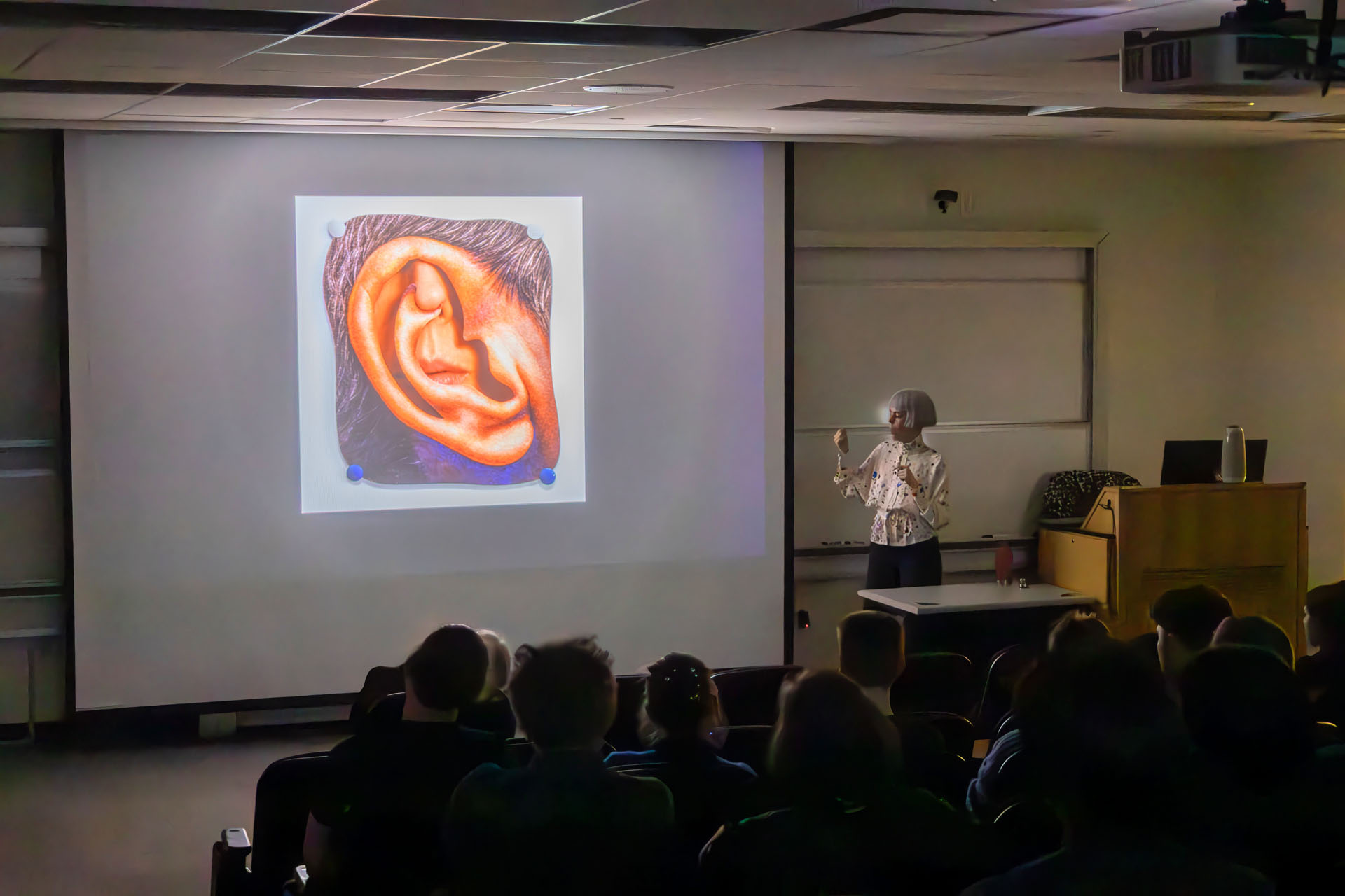 Colleen Asper, a woman with cropped white hair, speaks in front of a screen showing an enlarged crop of an ear.